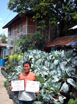 Blasa Perez holds her two certificates of recognition with  her  backyard vegetable garden behind her. (CDN Photo/Melissa Cabahug)