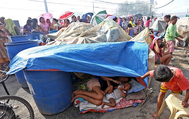 Victims of the fire that hit Barangay Guizo in Mandaue City use tarpauline as their temporary shelter while others line up (background) in front of the barangay hall to get relief goods from the  City Social Welfare and Services. (CDN PHOTO/JUNJIE MENDOZA)
