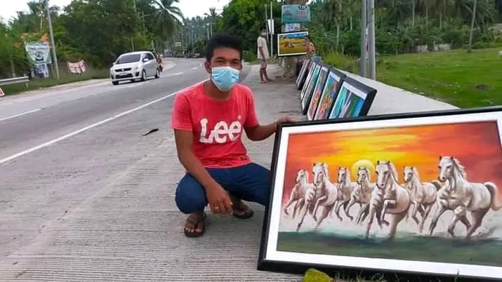 One of self-taught painter Salvacion's partners show one of the the painter's paintings displayed on the roadside in a town in northern Cebu. | Contributed photo