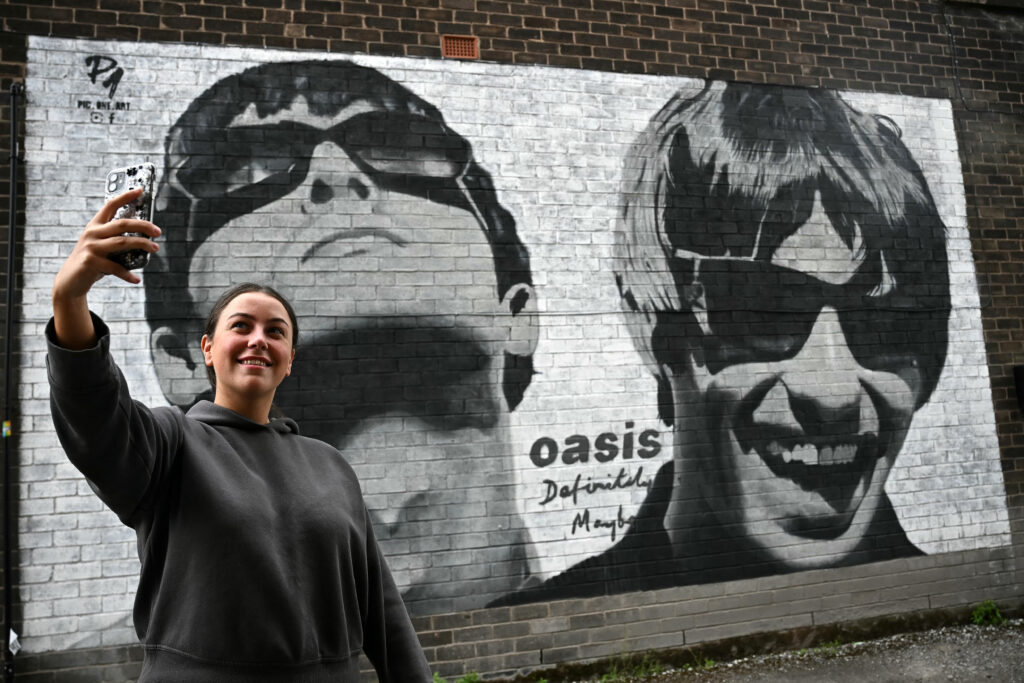 Oasis hometown abuzz after reunion tour announced. In photo is fan Emily McShane, takes a photograph of a new street artwork depicting Oasis' Liam and Noel Gallagher, created by Manchester-based street artist Pic.One.Art. on the side of the Sifters Record store in Burnage, a suburb of Manchester, northern England on August 27, 2024. | Photo by Paul ELLIS / AFP 