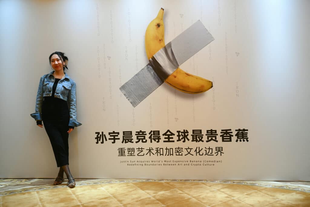 A woman poses for a photo in front of a poster depicting a banana artwork composed of a fresh banana stuck to a wall with duct tape before a press conference by Chinese-born crypto founder Justin Sun in Hong Kong on November 29, 2024. | Photo by Peter PARKS / AFP