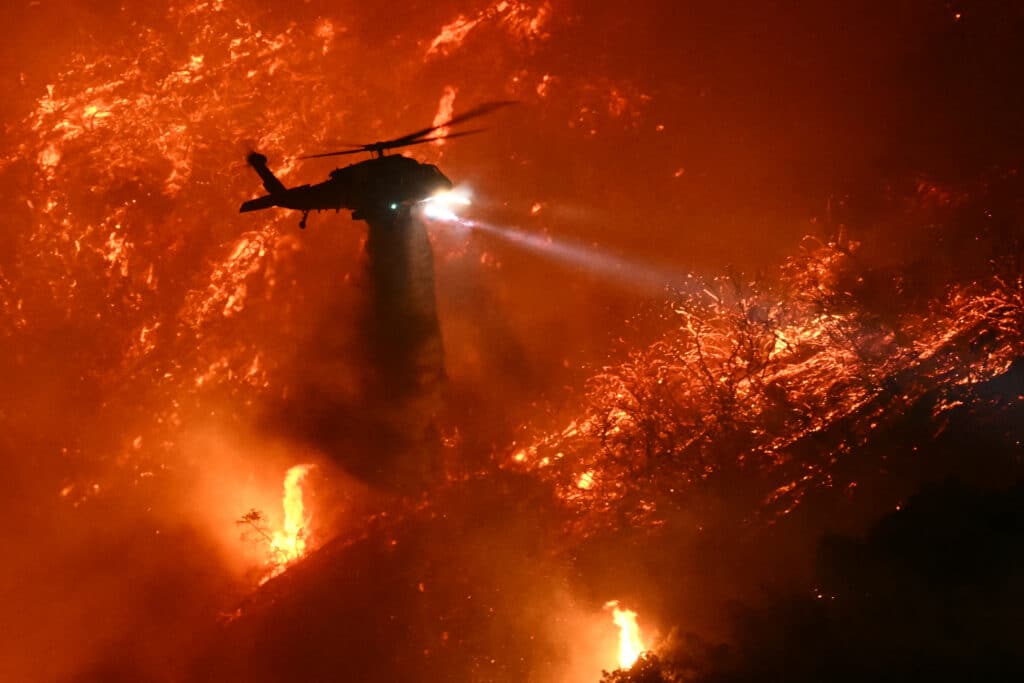 LA wildfires: 16 dead, 16 missing as fire crews try to corral blazes. A fire fighting helicopter drops water as the Palisades fire grows near the Mandeville Canyon neighborhood and Encino, California, on January 11, 2025. The Palisades Fire, the largest of the Los Angeles fires, spread toward previously untouched neighborhoods January 11, forcing new evacuations and dimming hopes that the disaster was coming under control. | Photo by Patrick T. Fallon / AFP