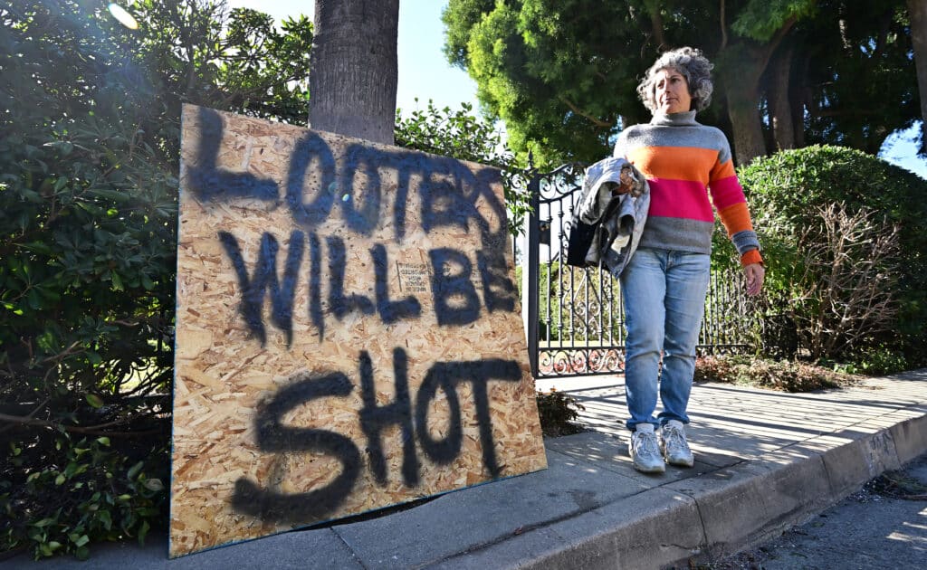 An Altadena resident walks past a sign in front of her home on January 13, 2025 warning looters they will be shot, which she put up due to concern of looters in the neighborhhood taking advantage of the situation following the Eaton Fire which caused widespread damage. | Photo by Frederic J. BROWN / AFP